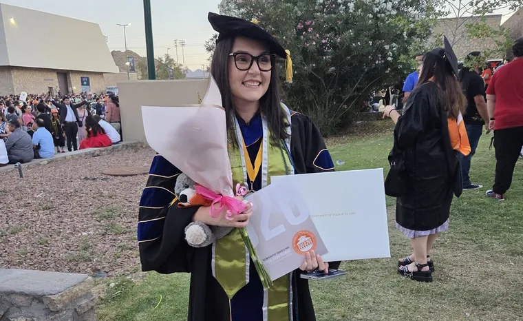 Karen Del Rio Guzman in doctoral regalia holding her diploma at UTEP commencement.